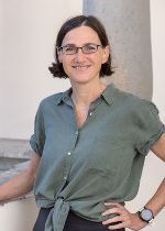 Dame mit kurzen dunklen Haaren und einer schmalen Brille in einer olivfarbenen Bluse vor einer Säule stehend.