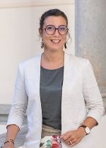 Eine Dame mit hellem Blazer und olivfarbenen Oberteil, dunklen zusammengemachten Haaren und einer Brille