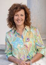 Frau mit rötlichen Locken, bunt gemusterten Kleid und roter Jacke vor hellgrauem Hintergrund