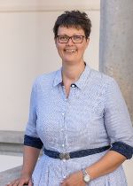 Hintergrund hellgrau, im Vordergrund junge Frau mit dunklen kurzen Haaren und einer Brille, freundlich lächelnd mit blitzblauem Oberteil