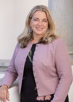 Frau in gemusterten Kleid und hellblauer Jeansjacke und langen blonden Haaren stehend vor hellgrauem Hintergrund