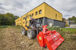 Gärtner auf einem Traktor sitzend. Am Traktor ist ein rotes Anbaugerät montiert. Er arbeitet auf einem Gemüsefeld, im Hintergrund ist ein gelbes Gebäude zu sehen - das Internat der Gartenbauschule.